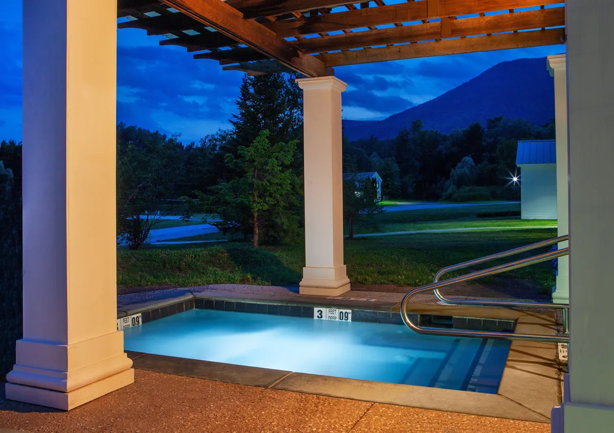 An illuminated outdoor pool under a wooden pergola, set against a breathtaking mountain backdrop. The area is softly lit for evening swims or stargazing, creating a magical and inviting nighttime atmosphere.