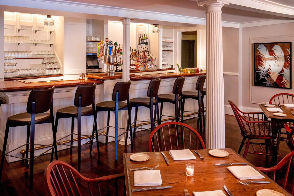 A cozy, stylish interior featuring a bustling bar counter with patrons socializing, dining tables set for meals, and eye-catching local artwork adorning the walls. The atmosphere is lively yet comfortable, highlighting gatherings over food and drinks.