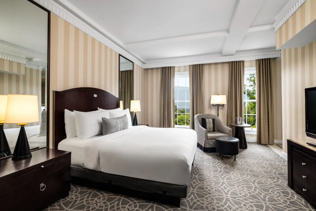The image shows a serene, sunlit studio interior at the Equinox Golf Resort & Spa in Manchester, Vermont. The inviting space features a plush bed beside a large window streaming in natural light, a cozy armchair perfectly positioned for enjoying the view or reading, a TV for evening relaxation, and elegant touches like a stylish lamp. The overall mood is calm, warm, and welcoming.