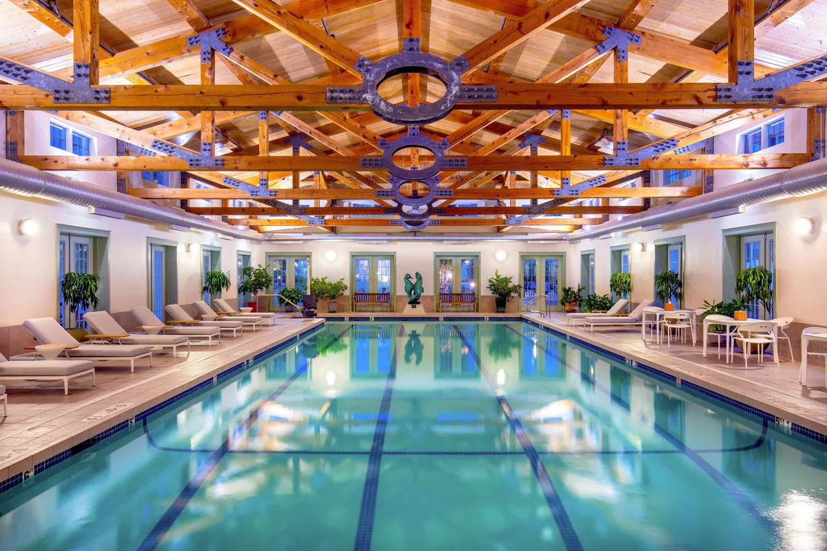 A tranquil and luxurious interior setting with a sparkling swimming pool framed by striking wooden beams, comfortable loungers, elegant sculptures, and a nearby dining area. The scene radiates peacefulness and invites relaxation, with opportunities for both swimming and social gatherings.