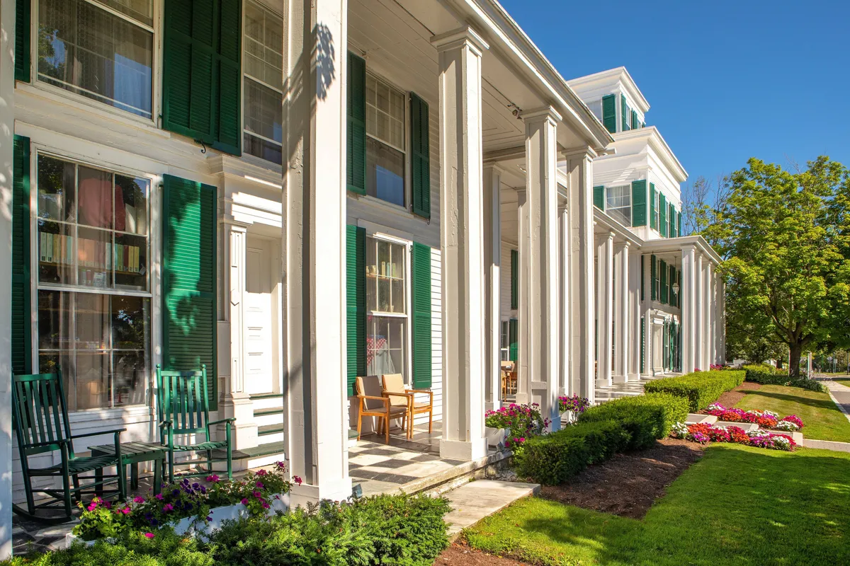This image showcases the inviting exterior of a Vermont resort, with charming green shutters, elegant white columns, cozy rocking chairs on a spacious porch, and meticulously maintained flower beds. The scene captures guests relaxing on the porch, enjoying the vibrant garden, and engaging in casual conversation, all set against the backdrop of the lush Green Mountains.