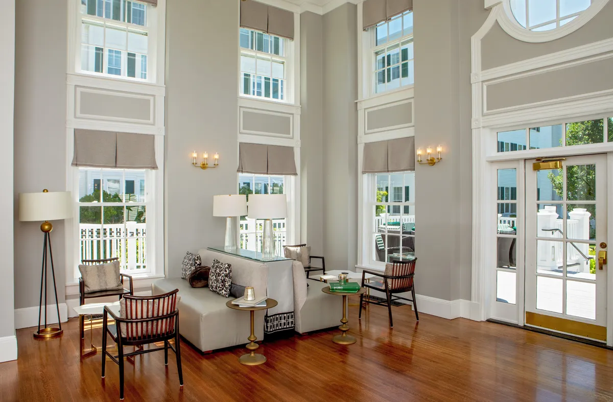A bright, welcoming studio interior featuring a comfortable seating area positioned near expansive windows. Sunlight pours through the glass, illuminating the modern, tasteful décor, plush textiles, and decorative accents on the tables. Stylish light fixtures and a serene atmosphere invite guests to relax and unwind.