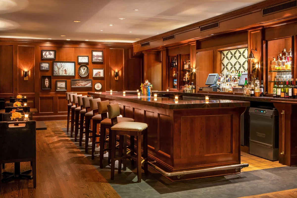 An elegantly designed interior bar lounge featuring a polished bar counter with wood paneling, stylish lighting fixtures, and sophisticated artwork on the walls. The space is filled with people socializing, enjoying drinks, and unwinding, evoking a luxurious yet welcoming atmosphere.