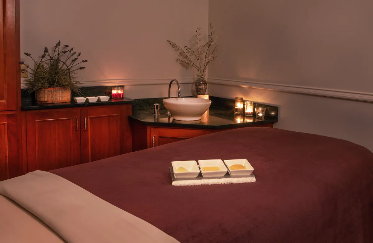 A tranquil spa room featuring a massage table adorned with fresh linens, wellness products arranged in decorative bowls, soft candlelight, and a touch of greenery from a decorative plant. The space radiates calm and invites guests to indulge in relaxing spa treatments or rejuvenating facials.